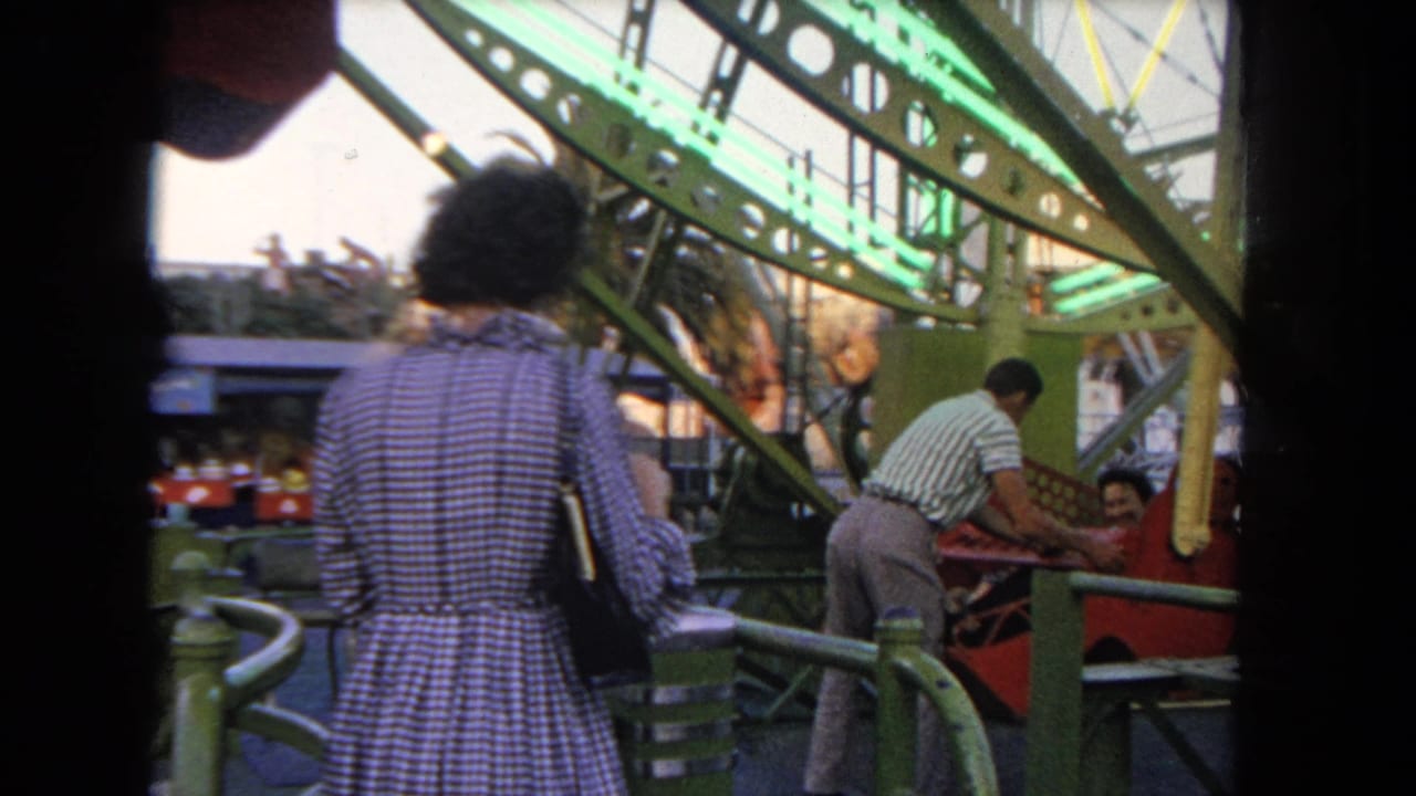 1962 Los Angeles carnival scene with a woman in a checkered dress watching attendants help riders into a green-framed ride.
