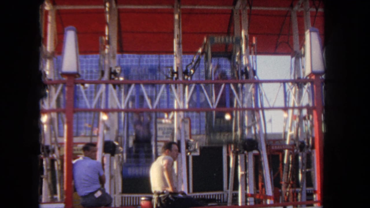 1962 Los Angeles home-movie frame showing two men watching a red vertical amusement ride with suspended cages and signs.