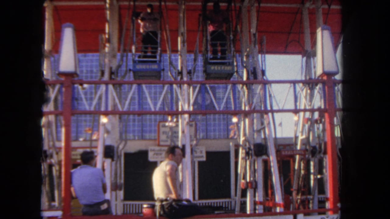 1962 Los Angeles frame with two men at the base of a red amusement ride as enclosed cars hang above them.