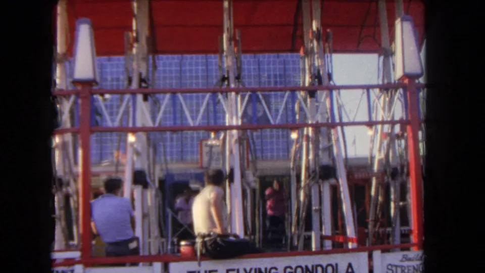 1962 Los Angeles home-movie frame showing two men below a red vertical ride, with partial gondola signage visible.