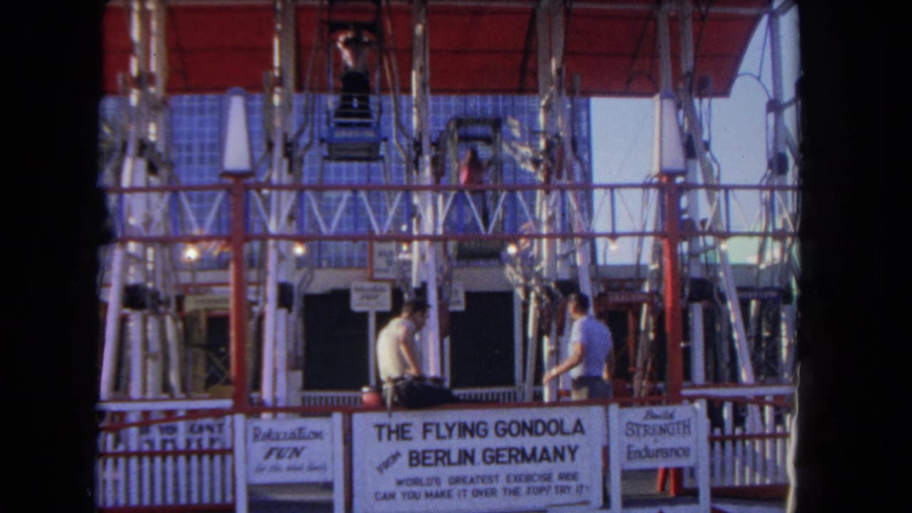 Two men stand by a Flying Gondola ride platform beneath a red canopy.