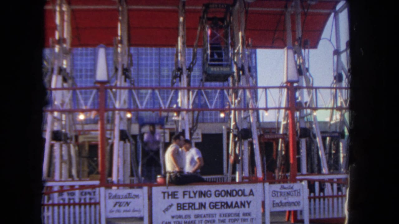 Two men stand near the ride entrance as a gondola hangs above them.