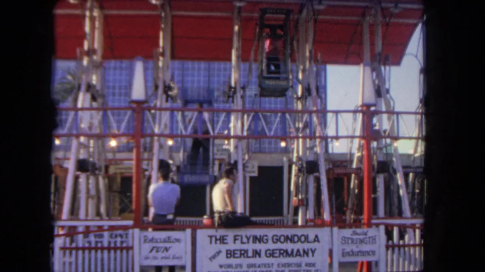 Two men watch the Flying Gondola ride structure from the platform area, with signage and suspended seats visible above.