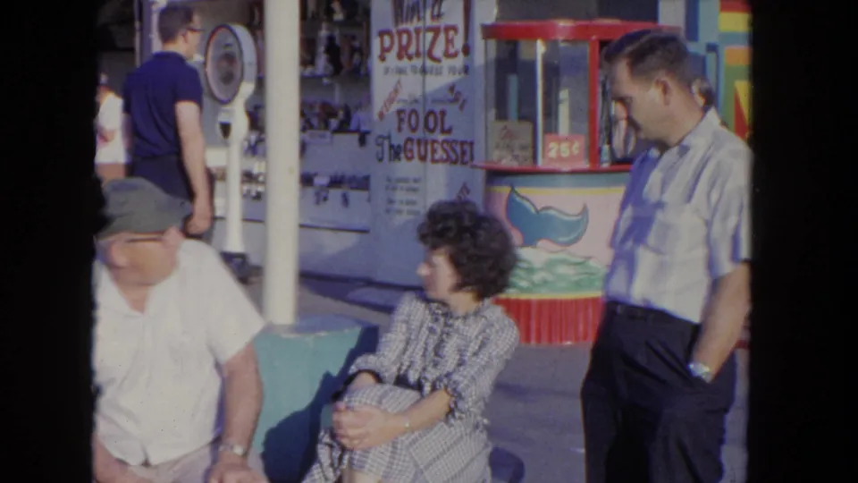 The 8mm frame shows a seated man in a cap, a seated woman in a checkered dress, and a standing man near a 1960s carnival game booth.