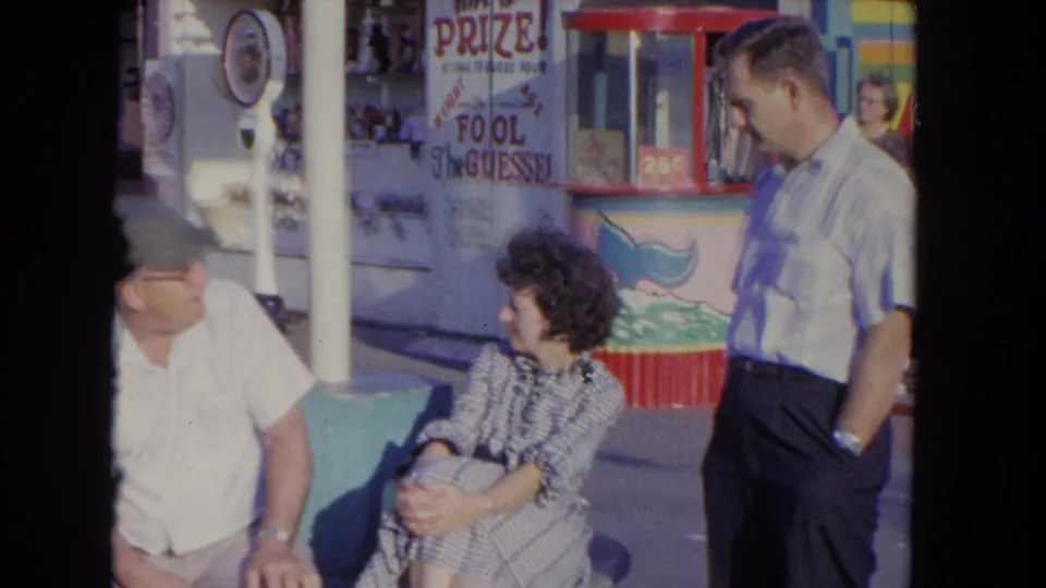 A 1962 Los Angeles home-movie frame shows adults conversing beside a carnival game booth with hand-painted prize signage and bright trim.