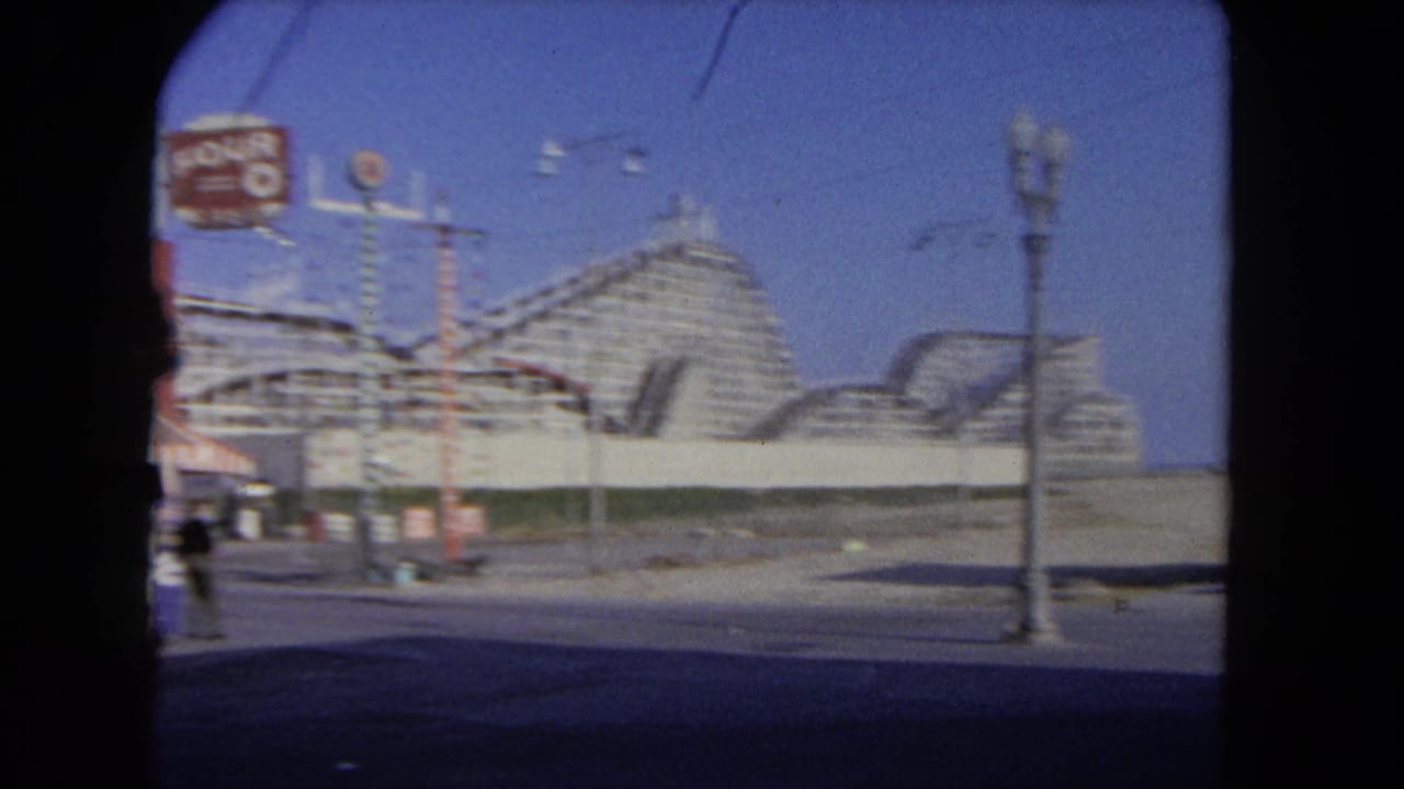1962 Los Angeles street view with a wooden roller coaster, overhead wires, lamps, and small pedestrians near the frame edge.