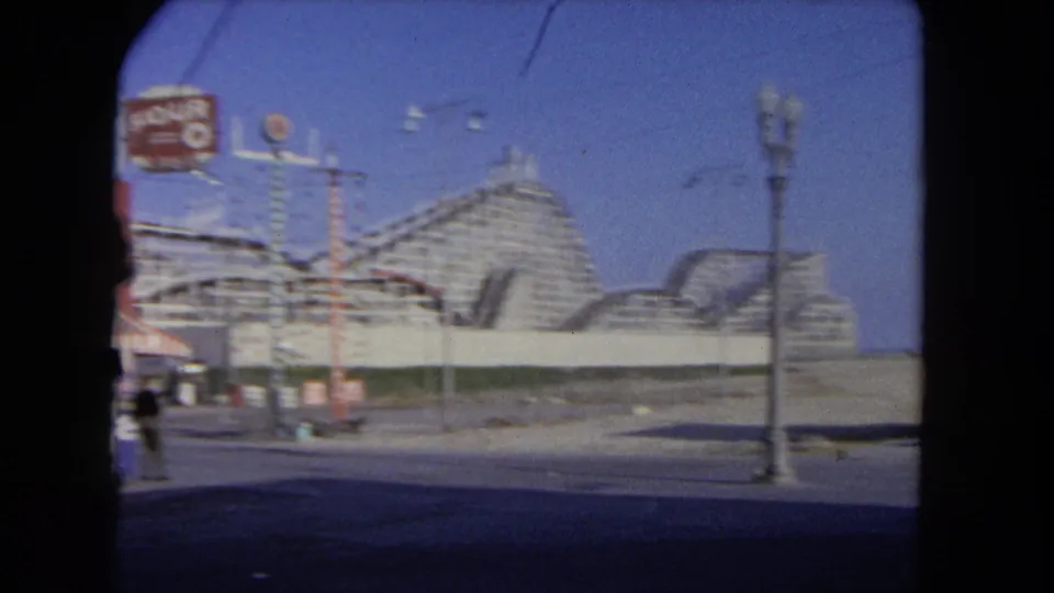 1962 Los Angeles street view with a wooden roller coaster, overhead wires, lamps, and small pedestrians near the frame edge.