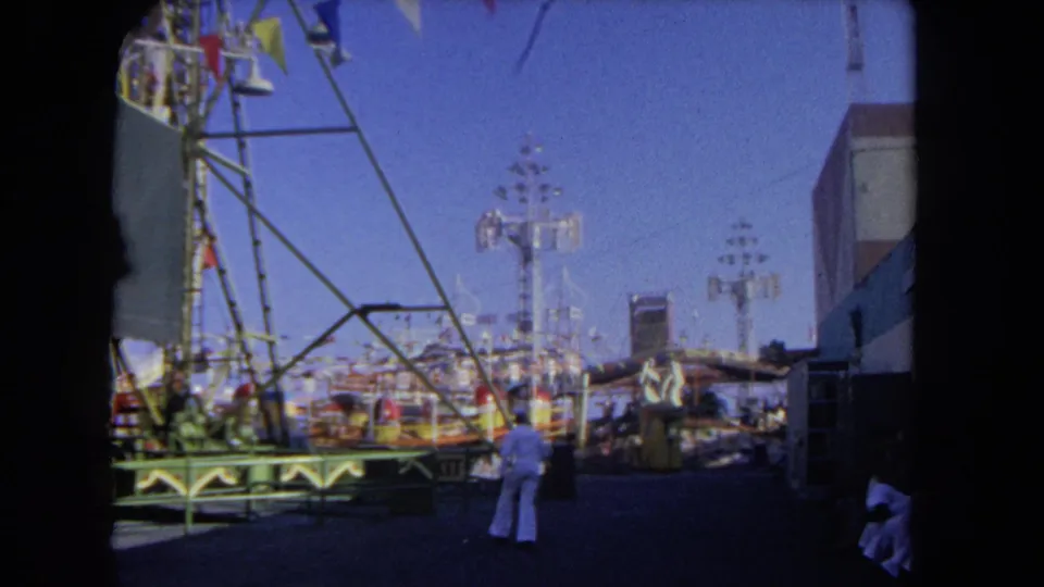 1962 Los Angeles amusement park view with a person in light clothing crossing the midway near painted railings, towers, and ride supports.
