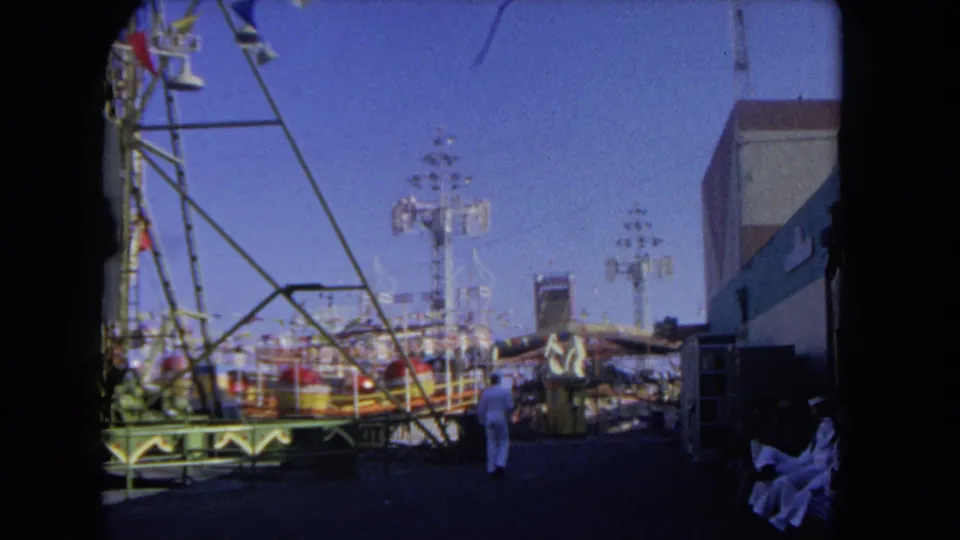 1962 Los Angeles amusement park frame showing seated visitors at the edge of the walkway and ride structures in bright daylight.