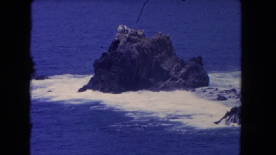 1962 Los Angeles-area 8mm frame showing foamy surf spreading beside a large coastal rock formation, with dark frame edges and aged film texture visible.