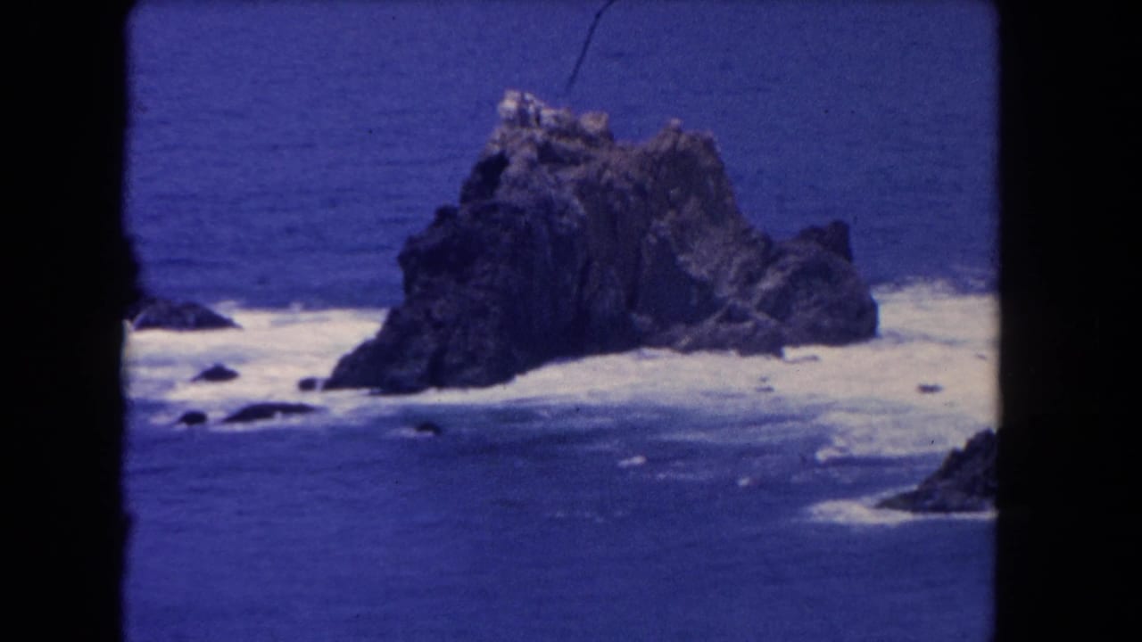 Large offshore rock with white foam spreading across the surrounding blue water.