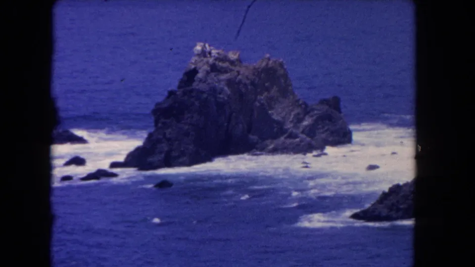 1962 Los Angeles-area 8mm frame showing the sea rock, nearby smaller rocks, and surf moving through blue water.