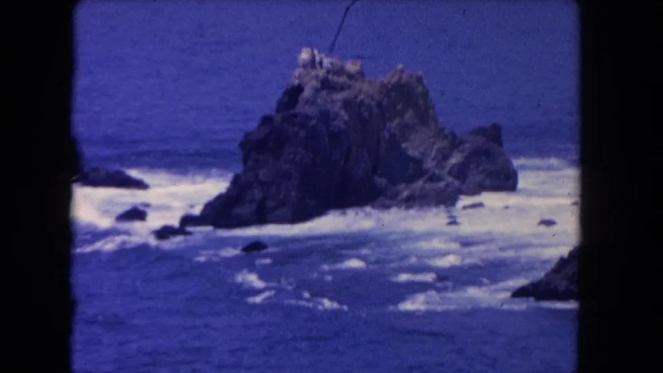 1962 Los Angeles-area coastal frame showing a jagged offshore rock with streaks of surf and visible 8mm borders.