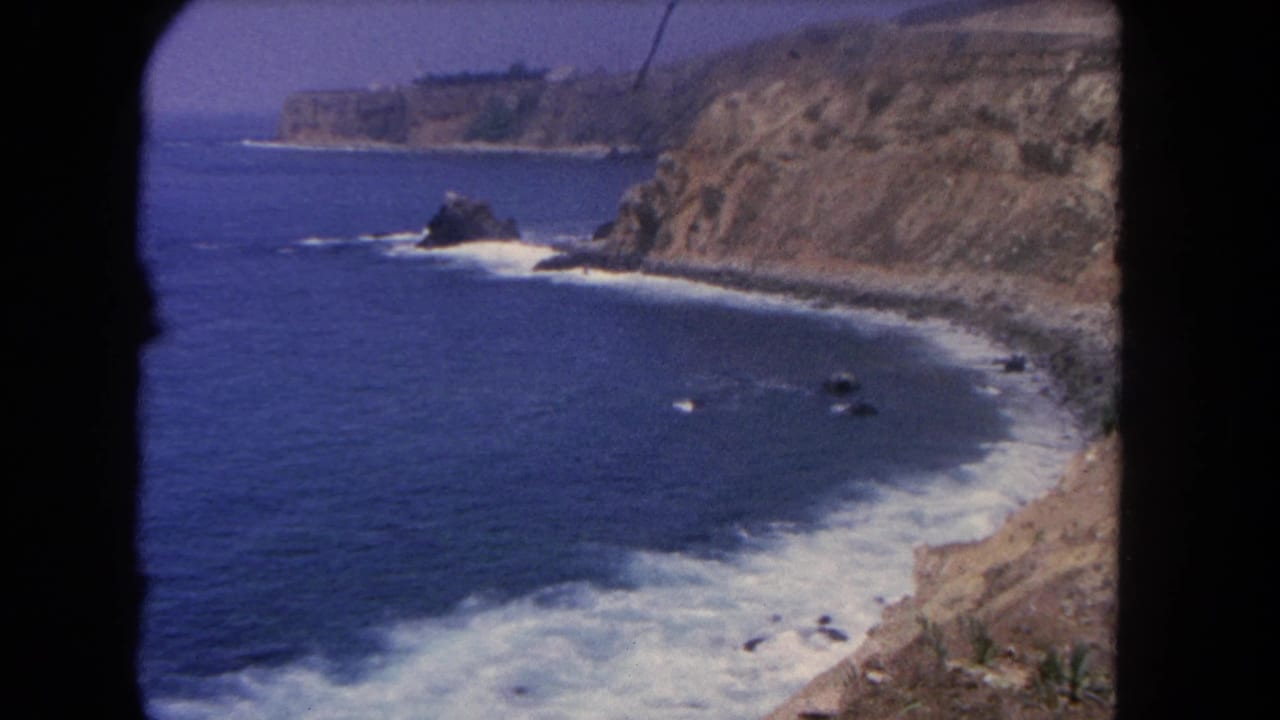 1962 Los Angeles-area coastal view with blue Pacific water, white surf, tan cliffs, and dark rocks seen through 8mm frame edges.
