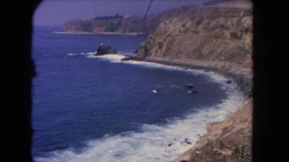 1962 Los Angeles-area coastal view with blue Pacific water, white surf, tan cliffs, and dark rocks seen through 8mm frame edges.