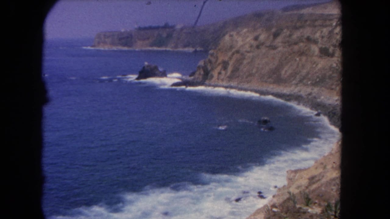 Los Angeles coastal home-movie frame showing blue water, foaming surf, tan cliff faces, and offshore rocks in a 1962 8mm scan.