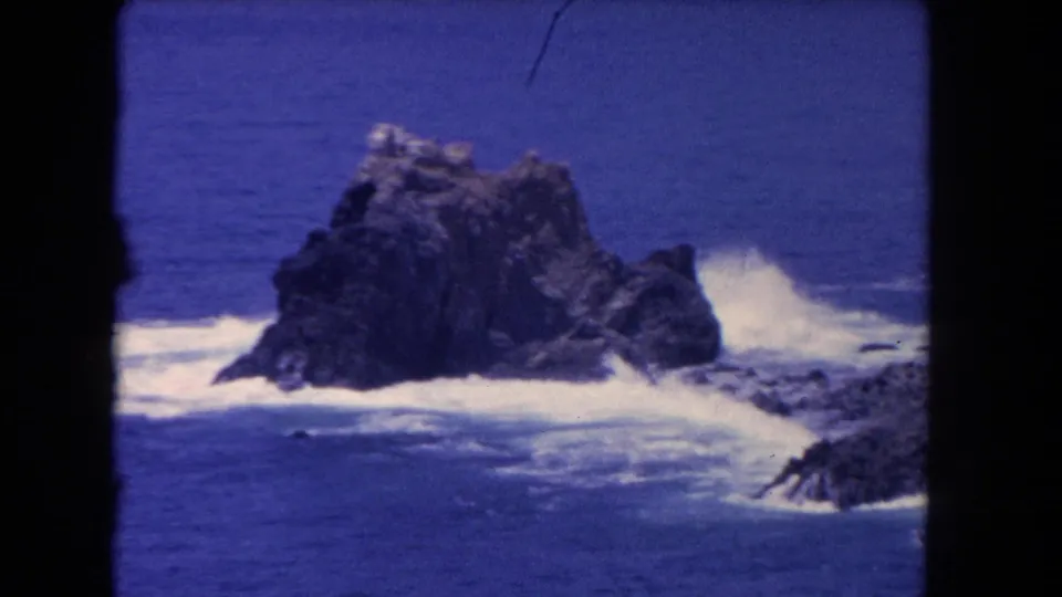 Closer 1962 coastal shot of a dark offshore rock surrounded by white surf and deep blue water, with soft 8mm color grain.