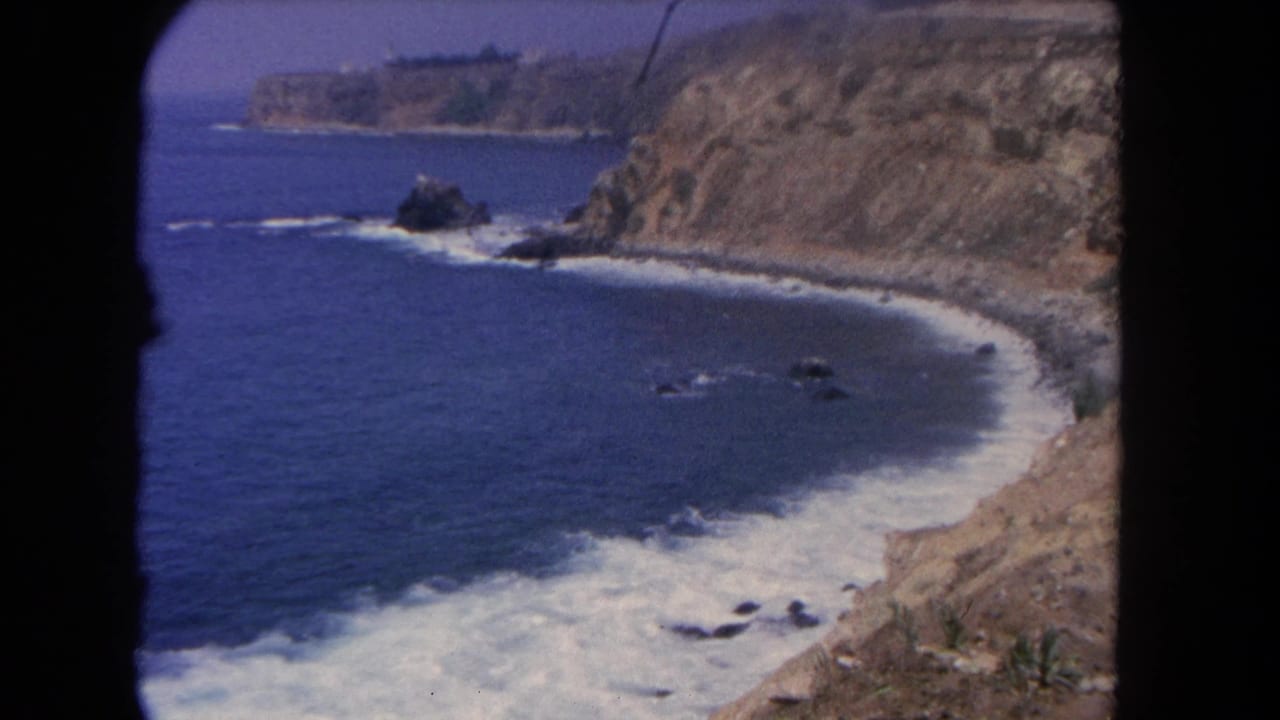 1962 Los Angeles shoreline seen from a cliffside viewpoint, with surf tracing the beach and rocks breaking the water surface.