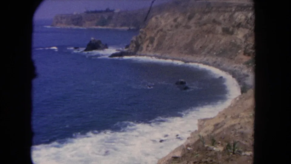 The final 1962 8mm frame cuts to a rugged California coastline with cliffs, surf, rocks, and a small curved beach.