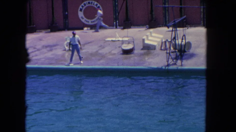 Home-movie frame of the Marineland stage with a trainer by the pool, visible props, and a dolphin partly out of view.