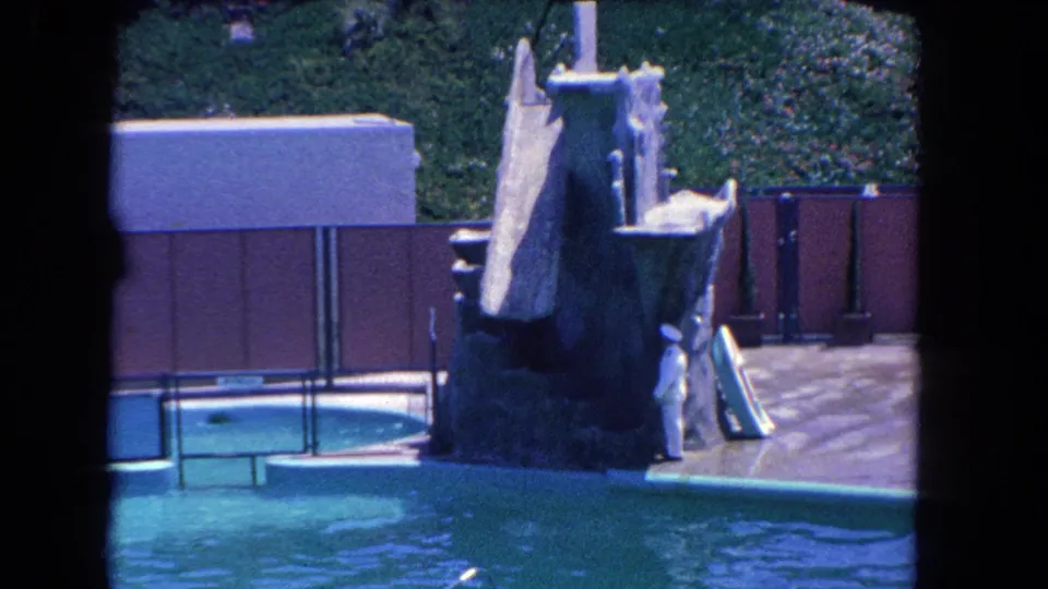 1962 Los Angeles 8mm frame showing a marine show platform, trainer, turquoise pool, brown fence, and hillside greenery.