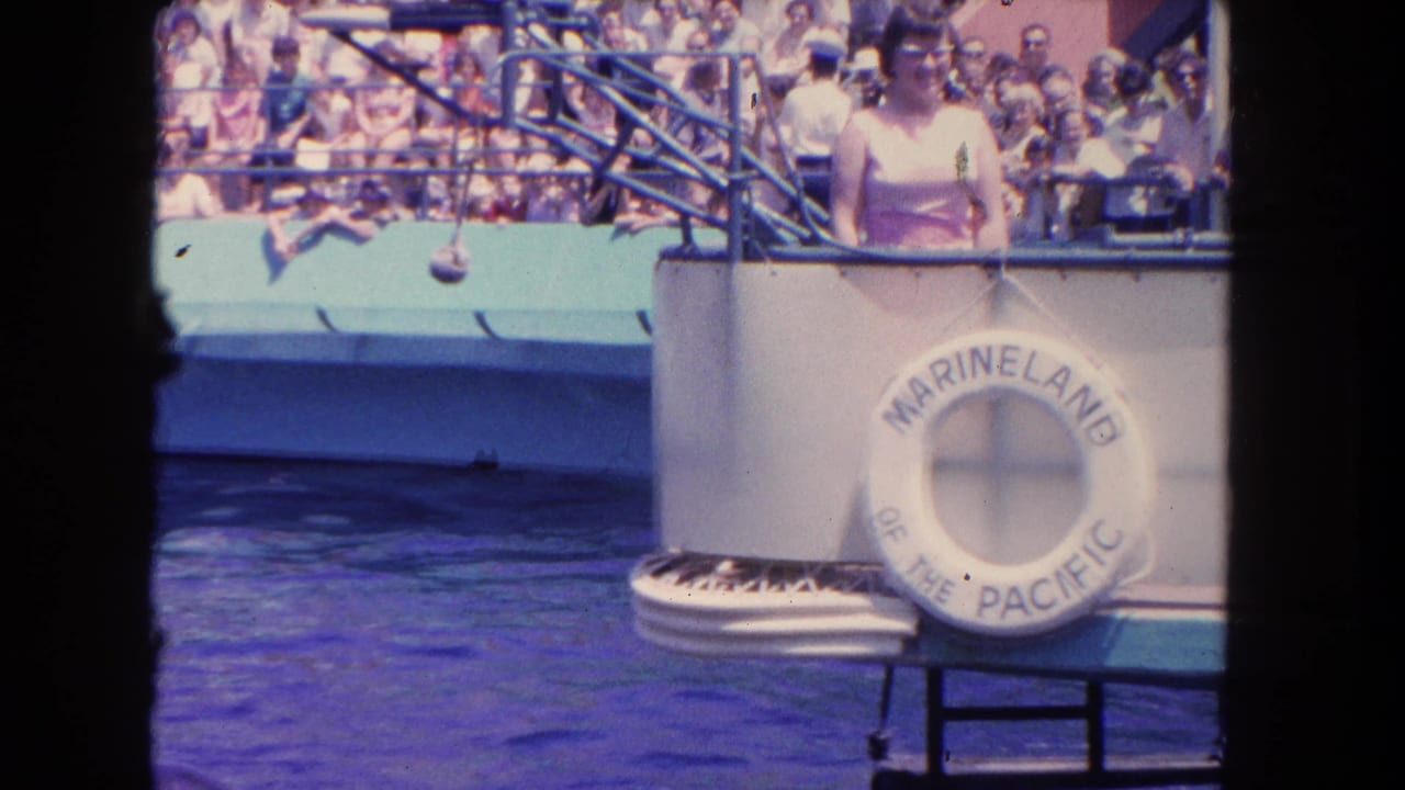 1962 home-movie frame at Marineland of the Pacific showing the pool platform, life ring, railings, and seated spectators.