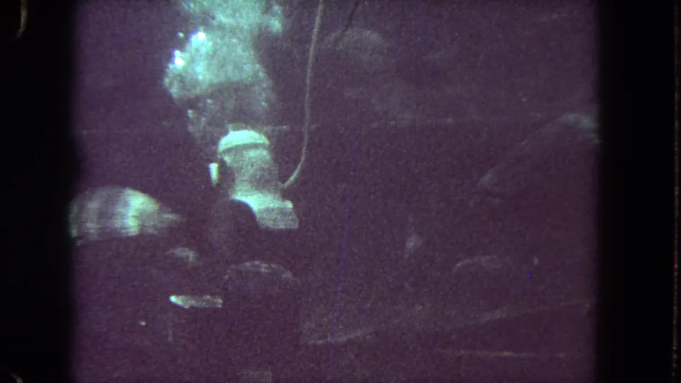 Helmeted diver visible from behind in a dim aquarium tank, with fish moving nearby in the 1962 Los Angeles footage.