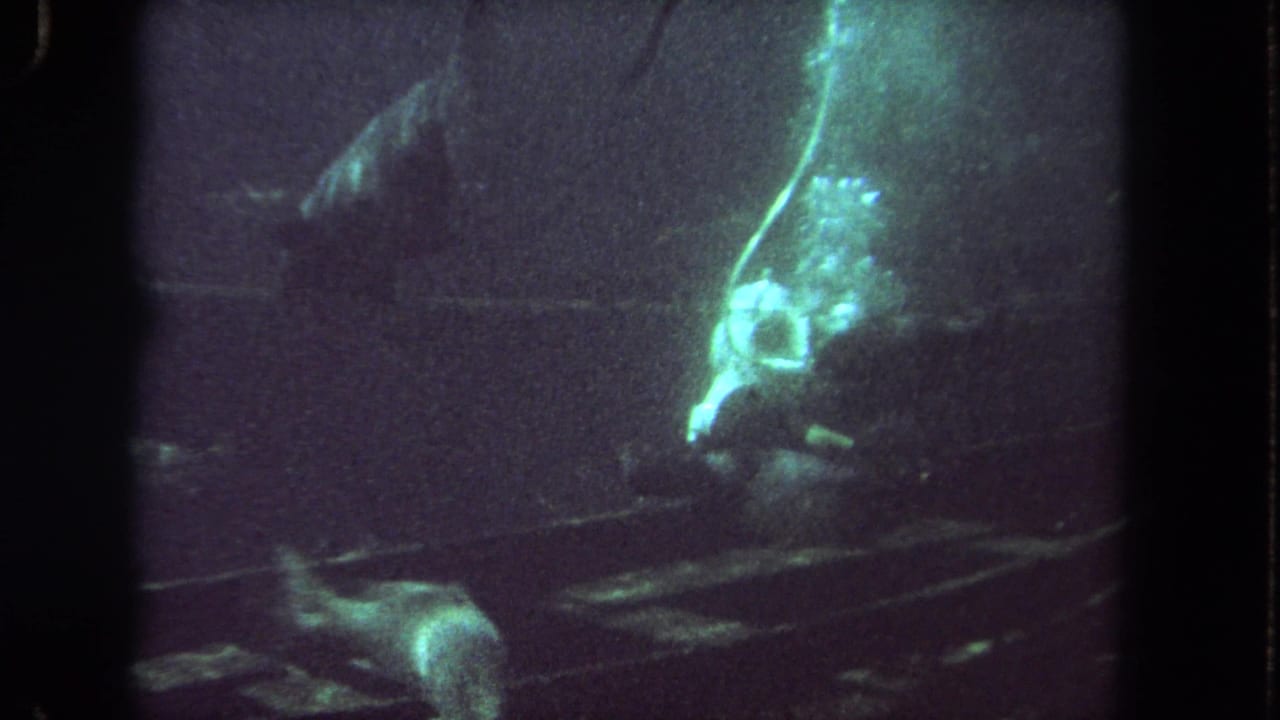 1962 Los Angeles 8mm frame showing a helmeted scuba diver near debris in a dim green underwater shipwreck-like setting.