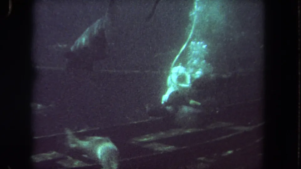1962 Los Angeles 8mm frame showing a helmeted scuba diver near debris in a dim green underwater shipwreck-like setting.