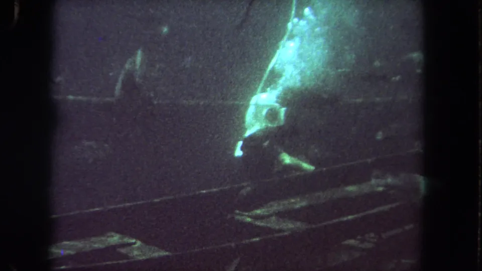 Helmeted diver appears to explore a submerged structure in 1962 Los Angeles 8mm footage, lit by a greenish underwater lamp.