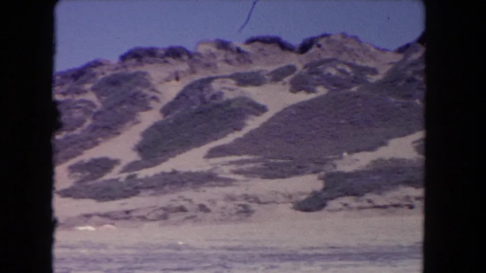 1962 Los Angeles-area 8mm view of dune hills with scrub, pale sand channels, and a soft foreground strip of open sand.