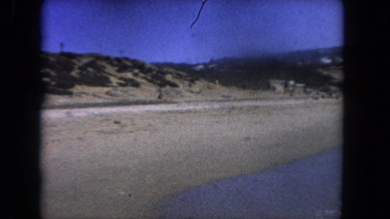 Wide view of sandy dunes and shoreline under a blue sky, framed by black film edges.