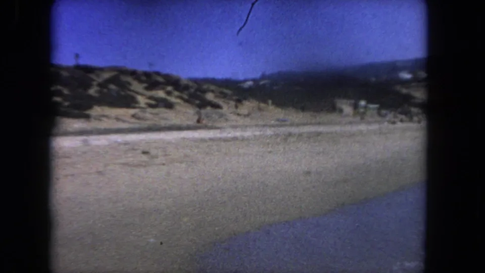 1962 8mm home-movie frame of a Los Angeles-area coastline with sand dunes, sparse scrub, water, and blue sky.