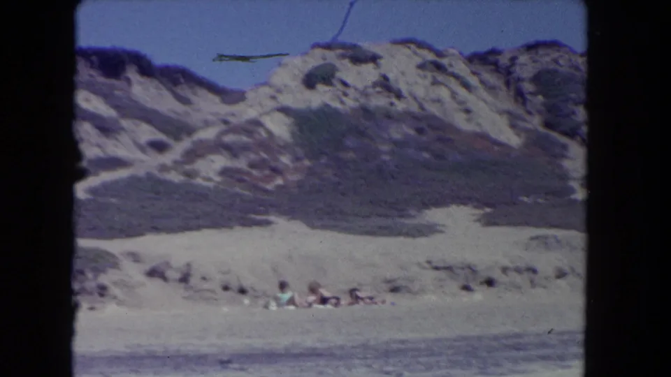 1962 8mm coastal frame showing a dune face, pale beach, water, and small distant people along the Los Angeles-area shore.