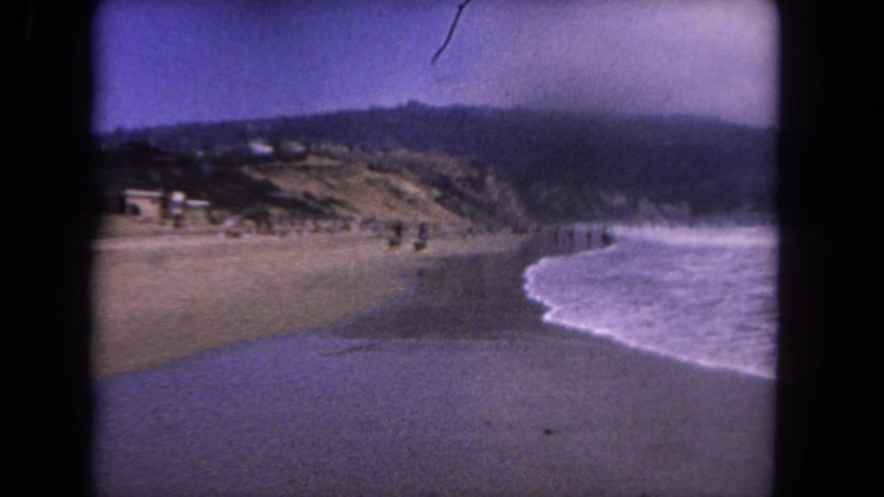 Wide sandy beach with waves washing in, distant people, coastal hills, and dark 8mm frame borders.