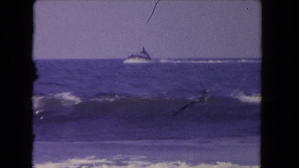 Distant speedboat and wake pass behind a darker foreground wave in purple-shifted 8mm Los Angeles beach footage from 1962.