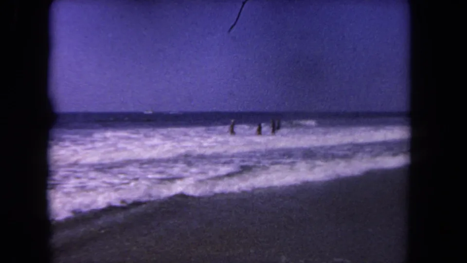 1962 Los Angeles beach home movie frame with small figures standing in rolling surf near the horizon.
