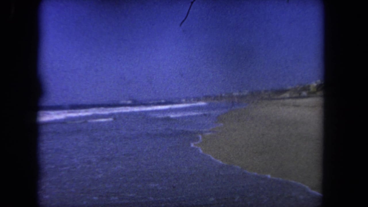 Sandy beach and ocean waves under a deep blue sky, framed by dark 8mm film edges.