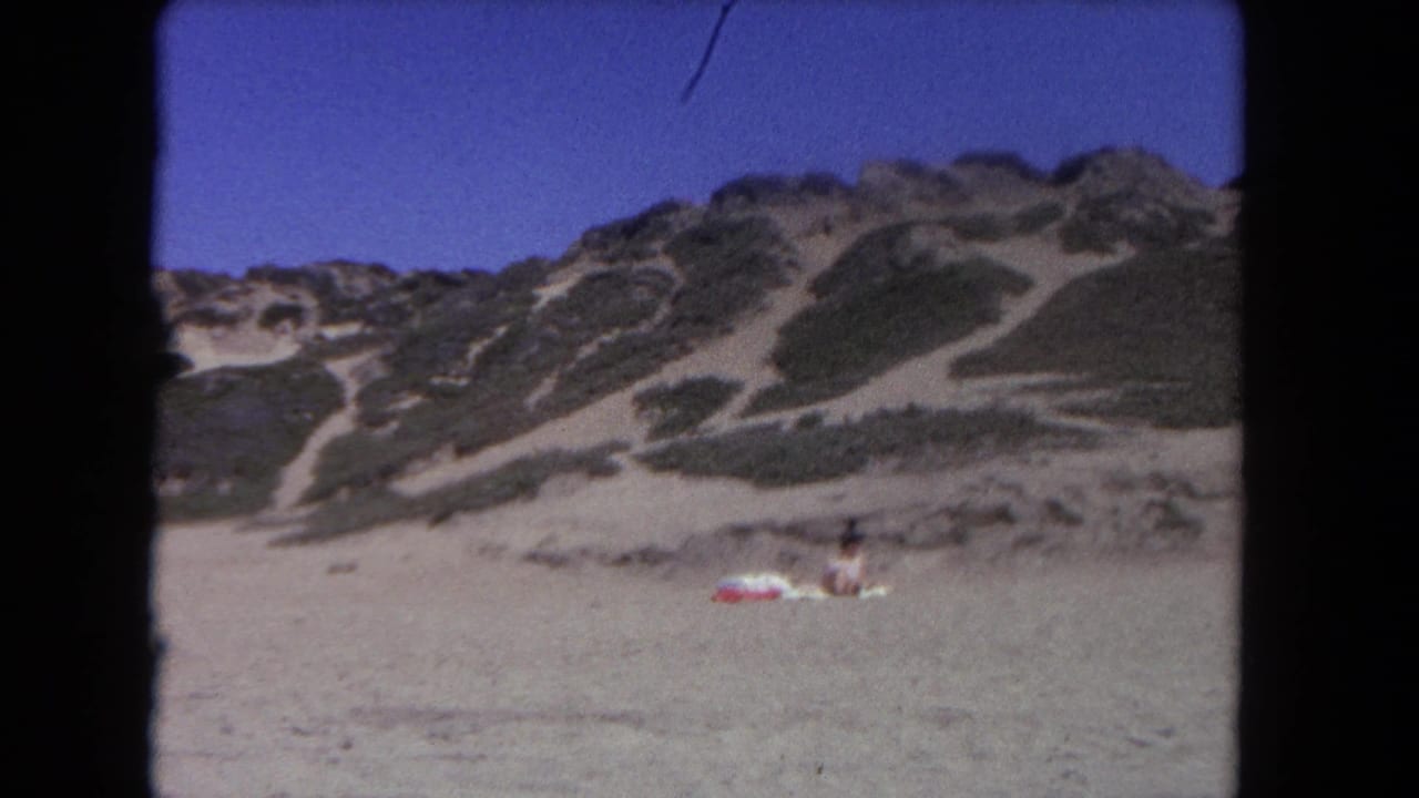 Distant person on a towel in front of sandy dunes with low vegetation and blue sky.