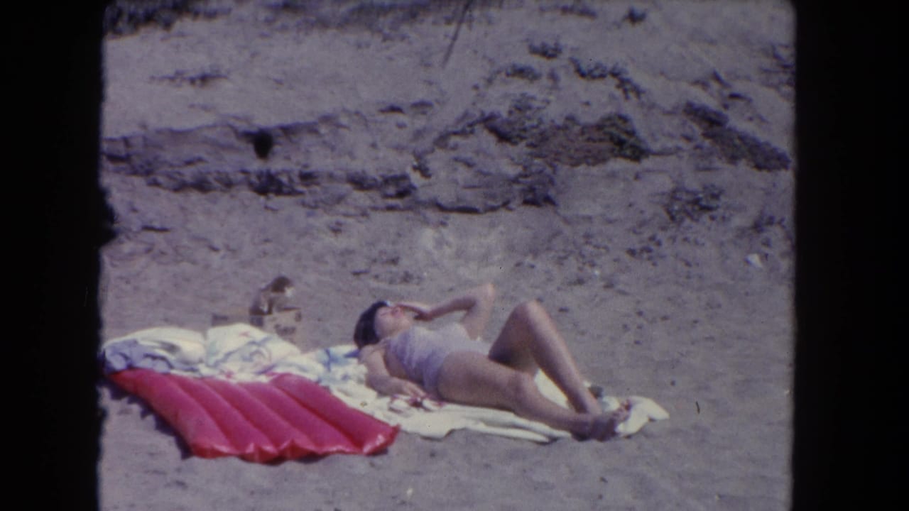 Woman sunbathes on a white towel beside a red inflatable float on a sandy beach.