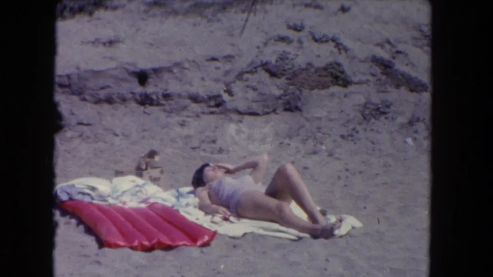 In 1962 Los Angeles home-movie footage, a woman in a light swimsuit lies on towels beside a red inflatable float on the sand.