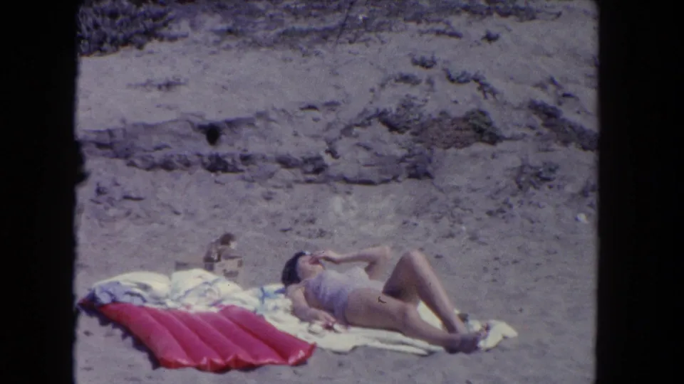 A woman in a light swimsuit reclines on towels in the 1962 Los Angeles beach scene, with the red float and sandy dune backdrop visible.