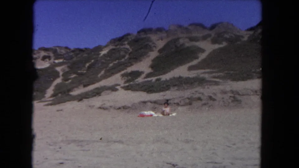 Wide 1962 Los Angeles beach view with the sunbather and red float small in frame below scrub-covered dunes and blue sky.