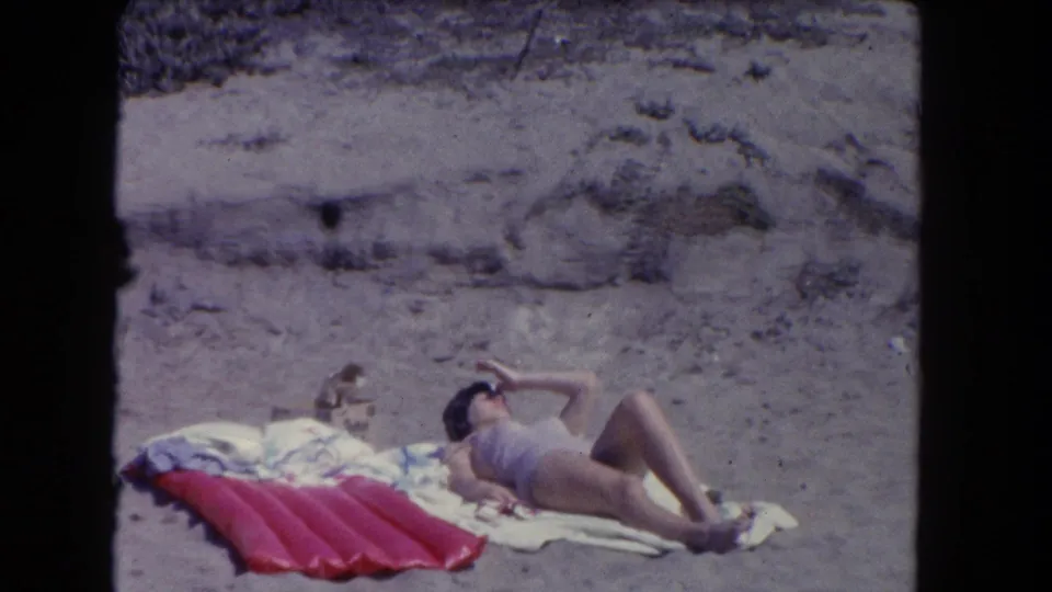 1962 Los Angeles beach footage showing a woman reclining on a towel beside a red inflatable mat on the sand.