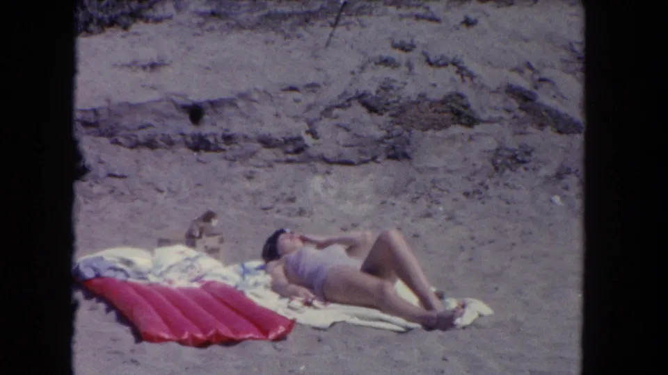 1962 Los Angeles beach scene with a woman resting on towels on the sand beside a red inflatable mat.