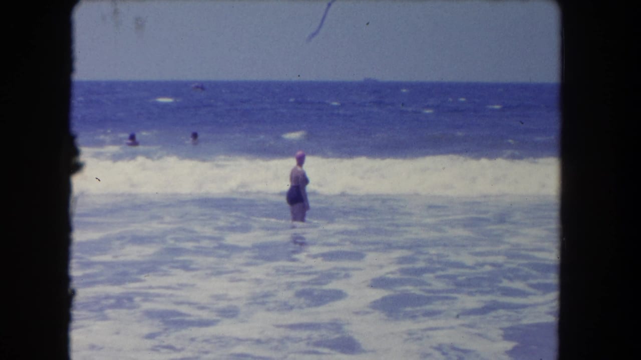 1962 Los Angeles-area beach footage showing a swimmer in a pink cap standing in foamy surf while other bathers appear farther out.