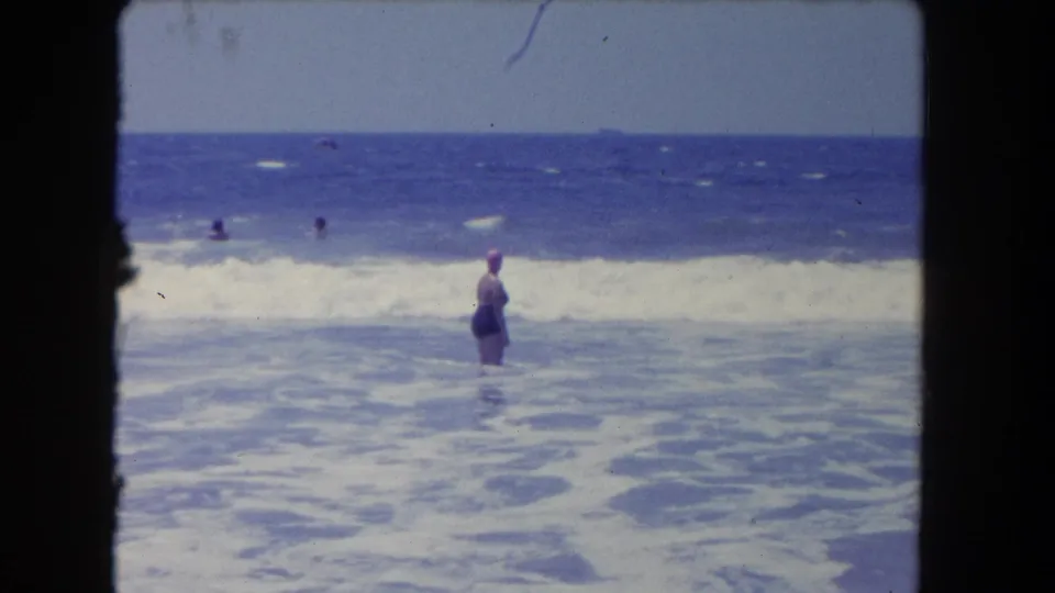 1962 Los Angeles-area beach footage showing a swimmer in a pink cap standing in foamy surf while other bathers appear farther out.