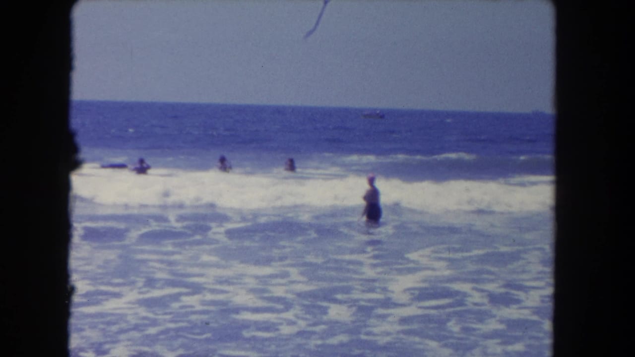 Woman in a pink swim cap stands in ocean surf with swimmers farther out.