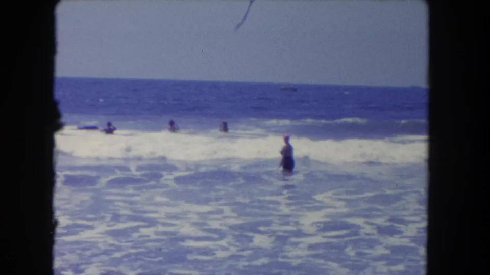 1962 Los Angeles beach home-movie frame showing a woman in a dark swimsuit and pink cap standing waist-deep in breaking waves.