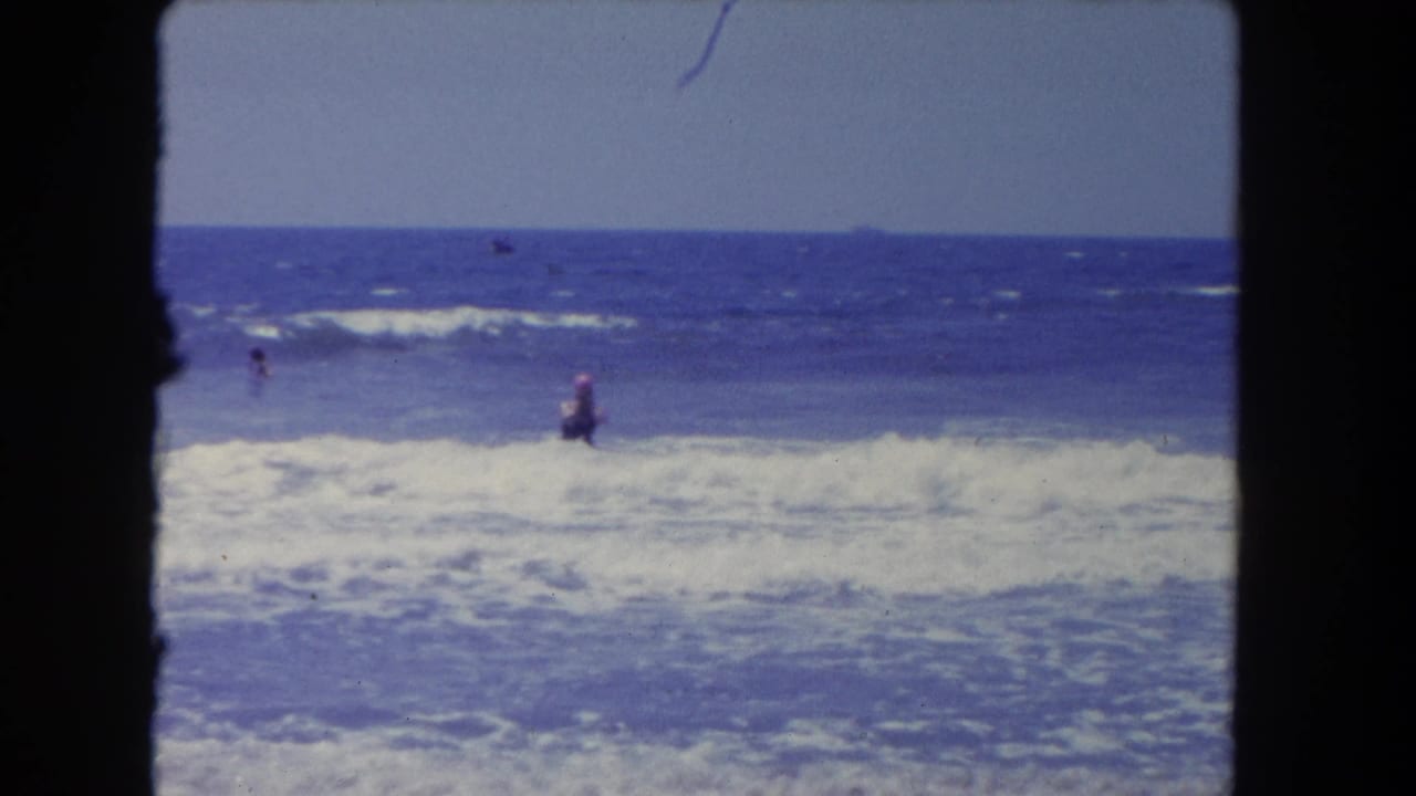 1962 Los Angeles 8mm frame showing a swimmer wading in white surf while other small figures appear farther offshore.